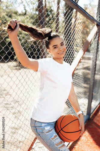 Brunette young woman with bright smile dressed in casual modern clothes posing with basketball at the stadium. © Screaghin