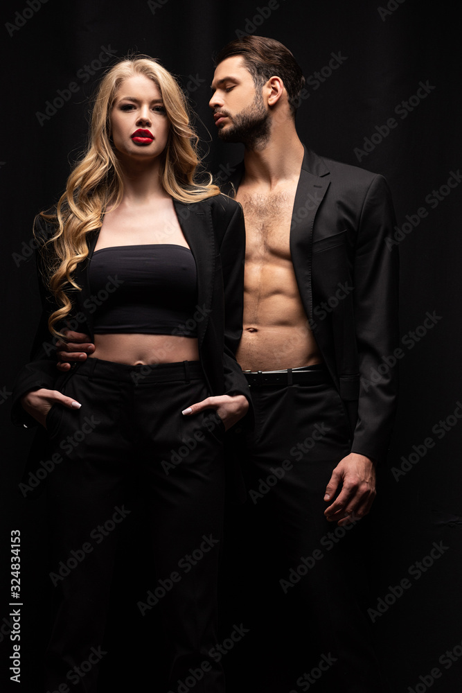 handsome man looking at stylish woman standing with hands in pockets isolated on black handsome man looking at stylish woman standing with hands in pockets isolated on black