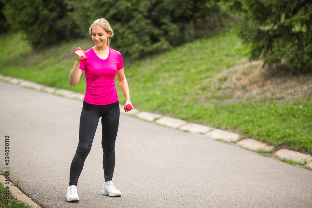 beautiful young woman in sports pink t-shirt with dumbbells in hands in the summer park beautiful young woman in sports pink t-shirt with dumbbells in hands in the summer park