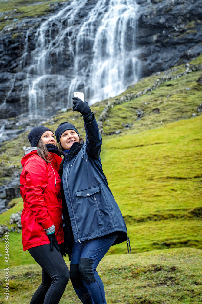 Two young female travelers taking selfie photos of herself surrounded with beautiful landscape of Faroe Island.