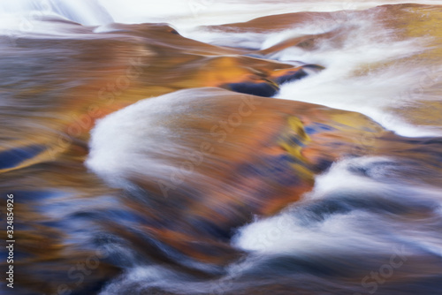 Landscape of a rapids at Bond Falls captured with motion blur and illuminated by reflected color from sunlit autumn foliage and blue sky overhead, Michigan's Upper Peninsula, USA