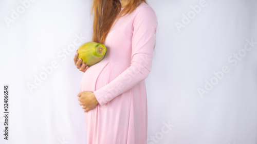 A close up photo of a pregnant young woman holding a papaya next to her belly showing the size of the baby in a beautiful dress. Photos of fetal growth at 22 weeks pregnancy.