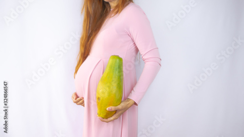 A close up photo of a pregnant young woman holding a papaya next to her belly showing the size of the baby in a beautiful dress. Photos of fetal growth at 22 weeks pregnancy. photo