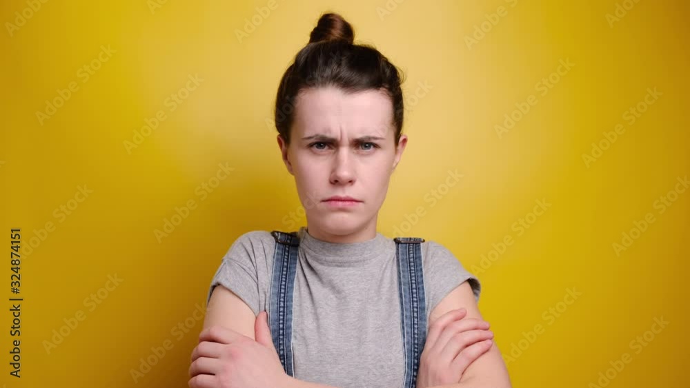 Distressed sad woman purses lips, looks with jealousy and regret, keeps hands crossed over chest, tells about unfair things, feels offended, wears grey t-shirt and denim, isolated on yellow wall Distressed sad woman purses lips, looks with jealousy and regret, keeps hands crossed over chest, tells about unfair things, feels offended, wears grey t-shirt and denim, isolated on yellow wall