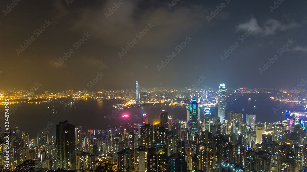 Hong Kong city skyline timelapse at night with Victoria Harbor and skyscrapers illuminated by lights over water viewed from mountain top.