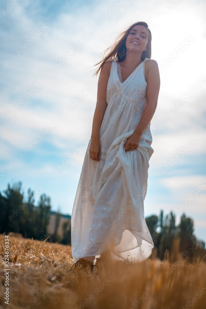 Fototapeta premium Rural lifestyle, a young caucasian blonde with a long white dress and a white hat in the dry wheat field