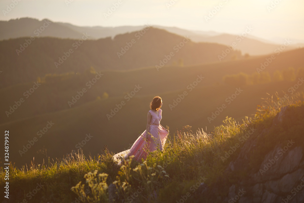 A girl in a pink dress with a long train dances in the sunset at the top of the mountain