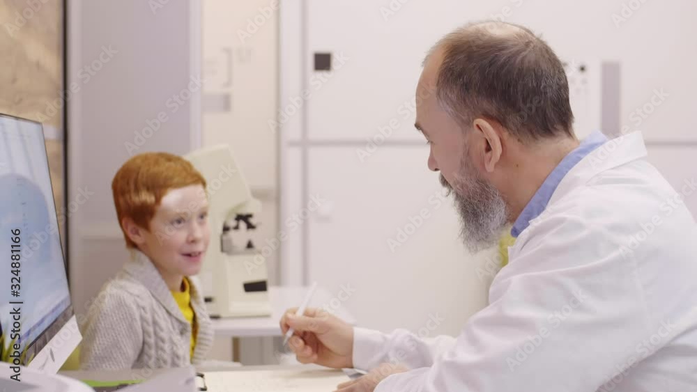Senior optometrist in lab coat sitting at desk, taking notes and talking with little redhead boy while giving medical consultation in clinic Senior optometrist in lab coat sitting at desk, taking notes and talking with little redhead boy while giving medical consultation in clinic