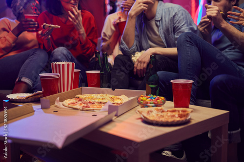 Close-up of pizza and drinks are on the table with young people sitting on sofa eating and drinking during a party