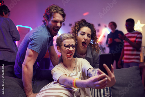 Group of friends making a face while posing at camera of mobile phone they making selfie portrait at a party © AnnaStills