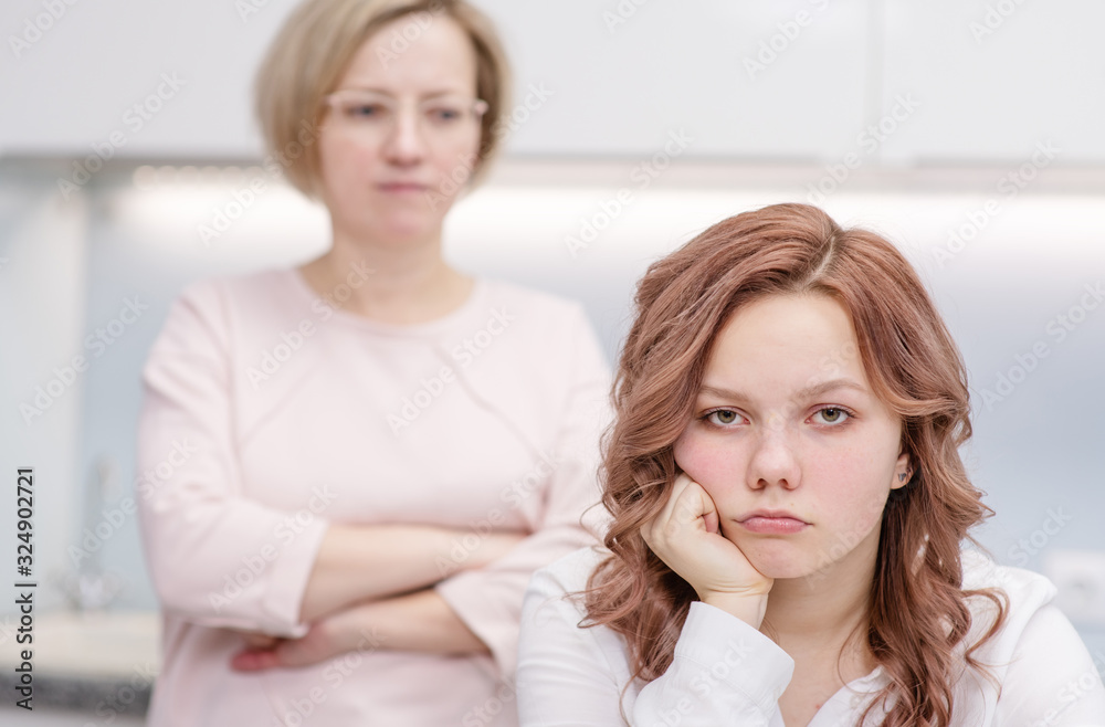 A teenage girl is sitting upset at a table in her kitchen. Her mother stands in the background, she is unhappy with her. Relationship problems, puberty A teenage girl is sitting upset at a table in her kitchen. Her mother stands in the background, she is unhappy with her. Relationship problems, puberty