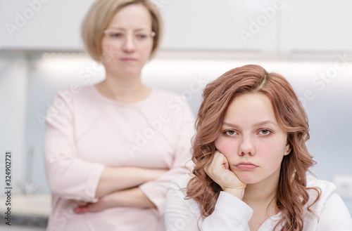 A teenage girl is sitting upset at a table in her kitchen. Her mother stands in the background, she is unhappy with her. Relationship problems, puberty
