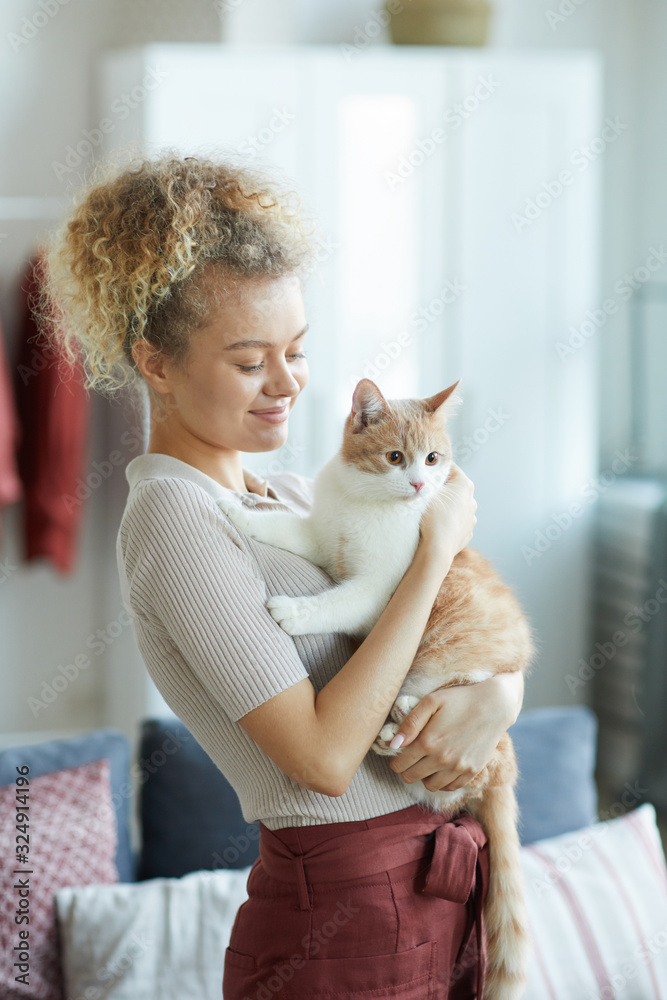 Young woman holding her domestic cat on her hands and loving it while standing in the room Young woman holding her domestic cat on her hands and loving it while standing in the room