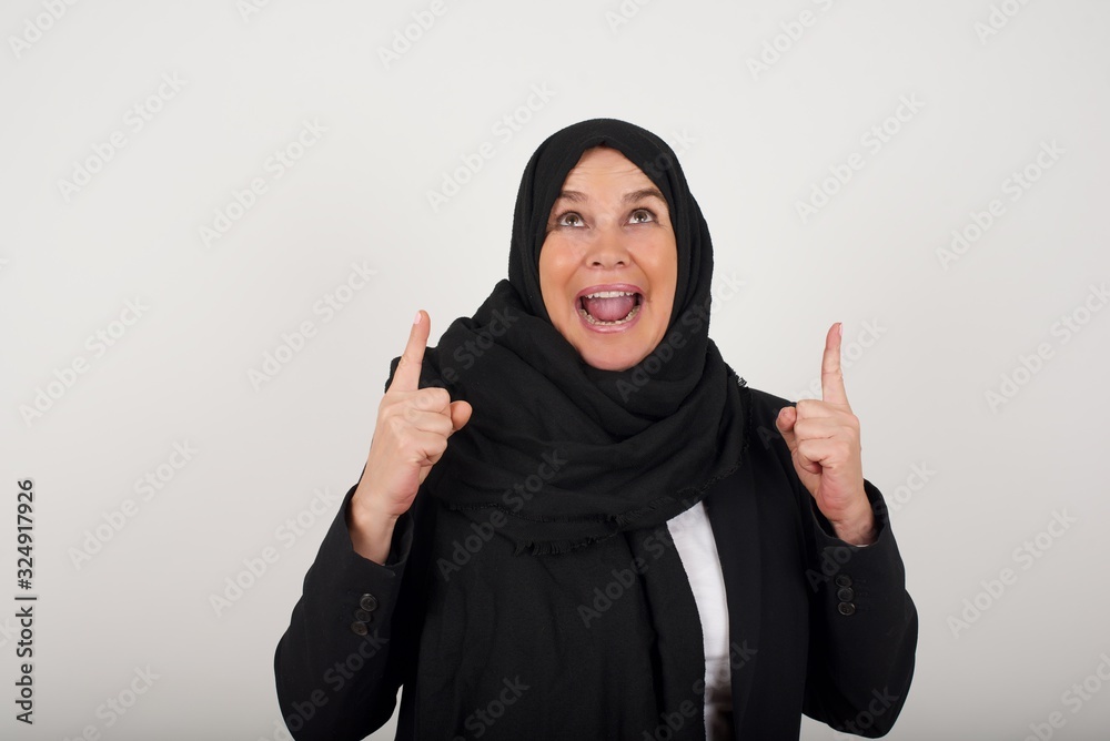 Charming carefree muslim woman with positive expression, points up with both index fingers, dressed in casual clothing, has broad interested smile, isolated over gray background. Look there, please. Charming carefree muslim woman with positive expression, points up with both index fingers, dressed in casual clothing, has broad interested smile, isolated over gray background. Look there, please.