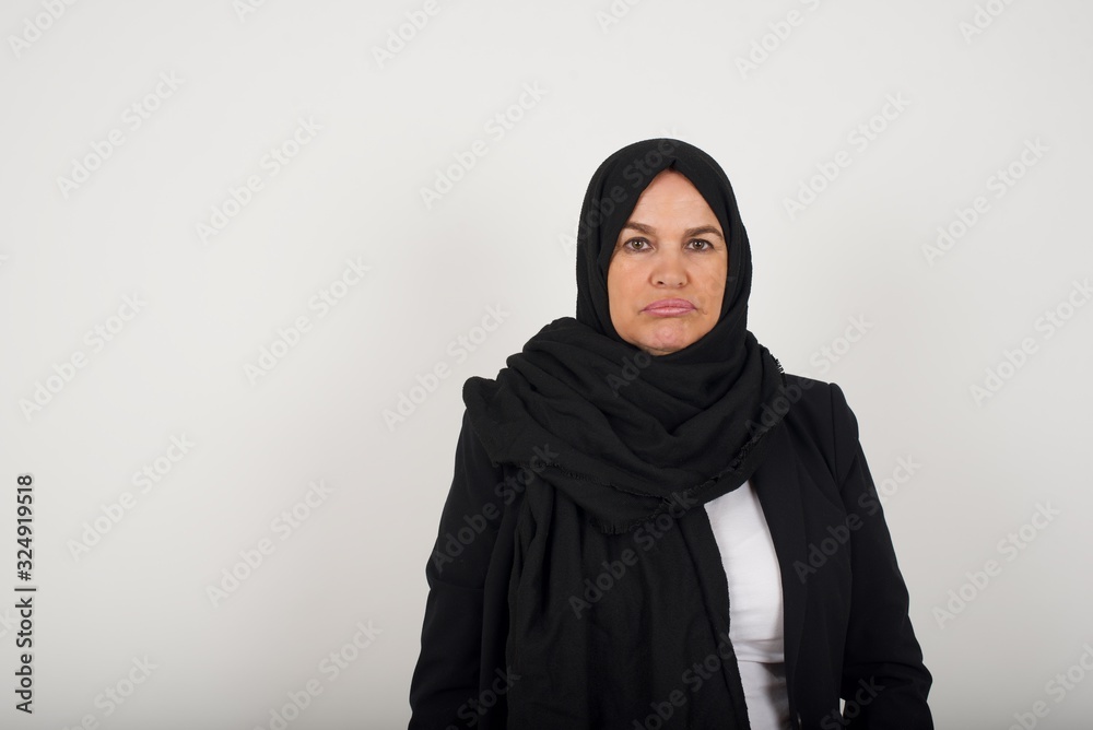 Gloomy, bored muslim lady frowns face looking up, being upset with so much talking hands down, feels tired and wants to leave. Angry sullen woman standing indoors. Gloomy, bored muslim lady frowns face looking up, being upset with so much talking hands down, feels tired and wants to leave. Angry sullen woman standing indoors.