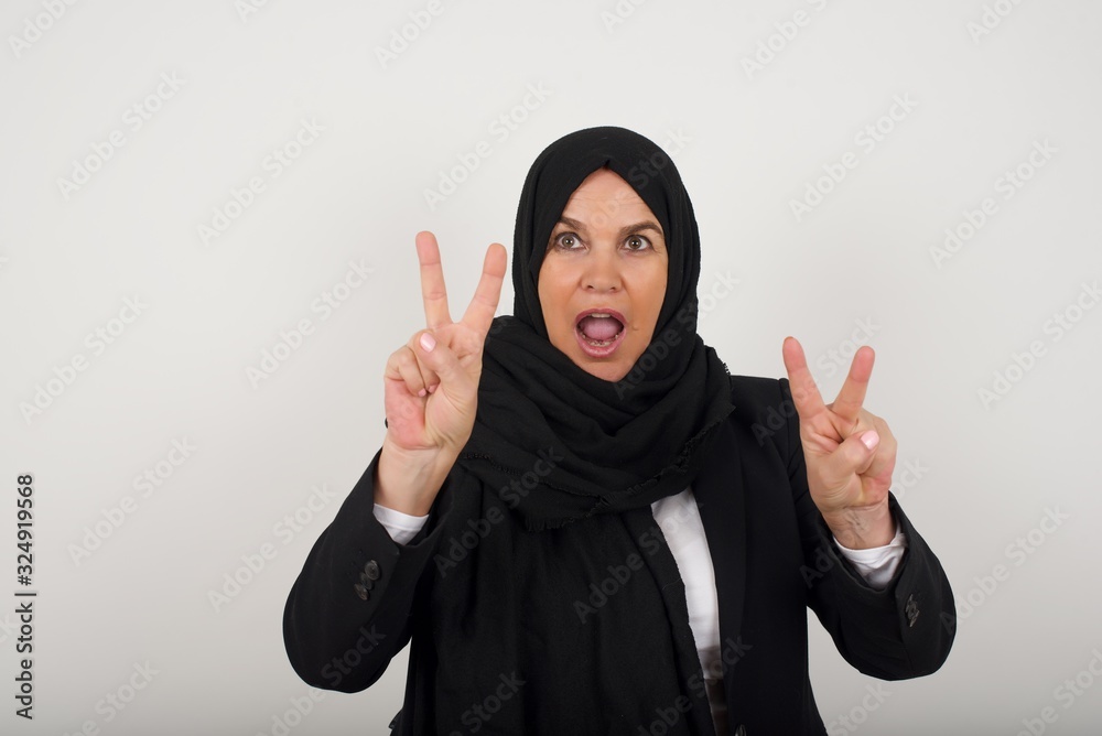 Isolated shot of cheerful muslim woman makes peace or victory sign with both hands, dressed in casual clothes, feels cool has toothy smile, isolated over gray background. People and body language. Isolated shot of cheerful muslim woman makes peace or victory sign with both hands, dressed in casual clothes, feels cool has toothy smile, isolated over gray background. People and body language.