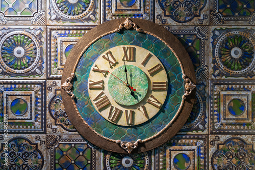 Ceramic wall clock with roman dial against the background of ancient fireplace tiles.