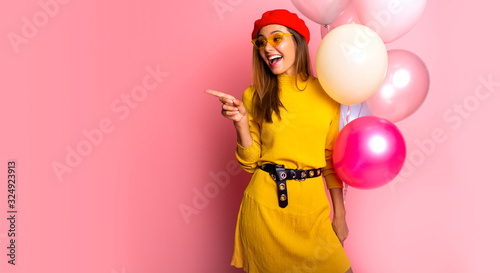 Young woman catching many balloons over isolated on pink background pointing finger to the side