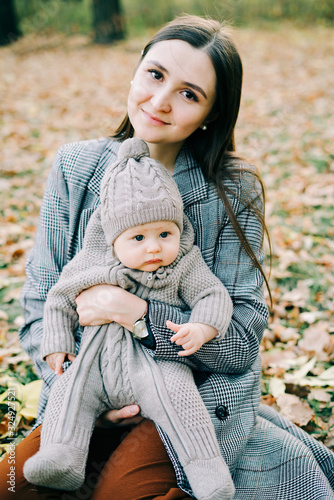 a young mother holds a cute little boy in a warm jumpsuit in the Park in a clearing strewn with autumn leaves, in the autumn time
