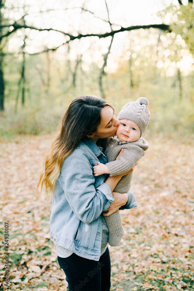 mom holds a little cute boy in a warm jumpsuit, kisses on the cheek in the Park in a clearing strewn with autumn leaves, in the autumn time mom holds a little cute boy in a warm jumpsuit, kisses on the cheek in the Park in a clearing strewn with autumn leaves, in the autumn time