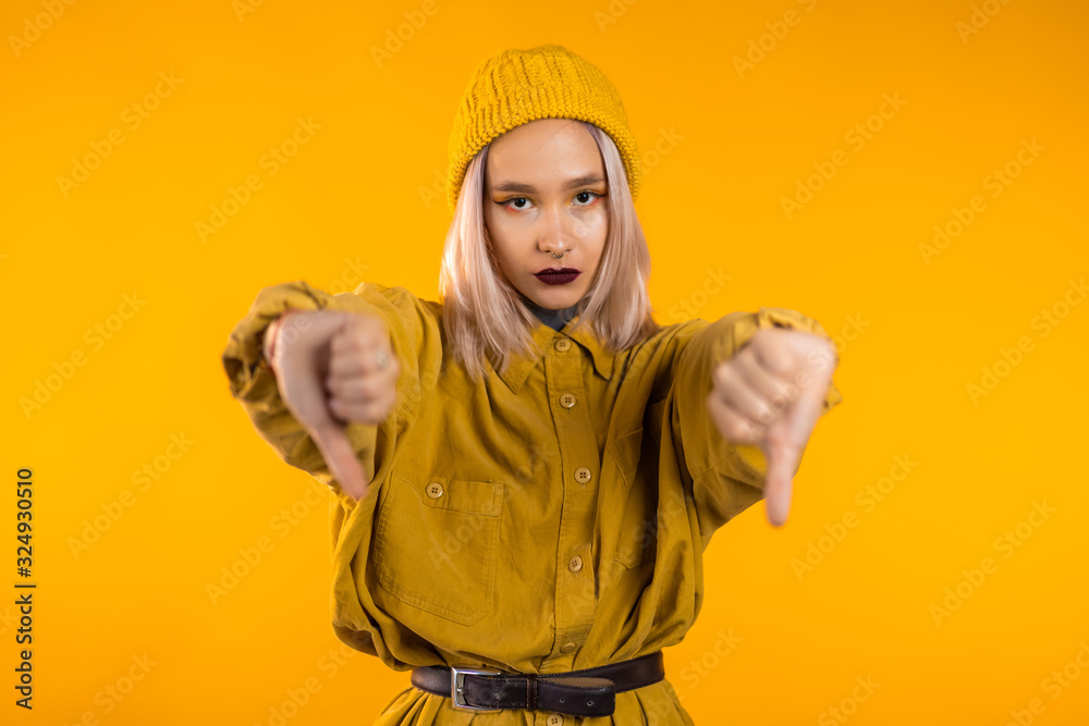 Young pretty woman standing on yellow studio background expressing discontent and showing thumb down gesture at camera. Portrait of girl with sign of dislike Young pretty woman standing on yellow studio background expressing discontent and showing thumb down gesture at camera. Portrait of girl with sign of dislike