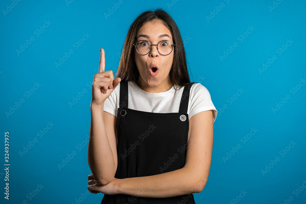 Portrait of young asian thinking pondering girl having idea moment pointing finger up on blue studio background. Smiling happy student showing eureka gesture. Portrait of young asian thinking pondering girl having idea moment pointing finger up on blue studio background. Smiling happy student showing eureka gesture.