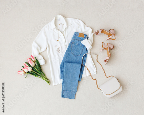 Blue jeans, white shirt, heeled sandals, bag with chain strap, jewelry, bouquet of pink tulips flowers on beige background. Women's stylish spring summer outfit. Trendy clothes. Flat lay, top view. photo