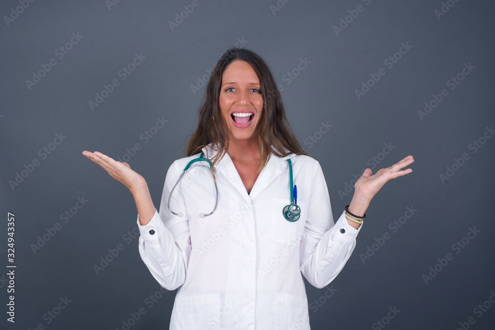 Portrait of beautiful doctor female looks with excitement at camera, keeps hands raised over head, notices something unexpected, isolated over gray wall. Lovely woman reacts on sudden news. Portrait of beautiful doctor female looks with excitement at camera, keeps hands raised over head, notices something unexpected, isolated over gray wall. Lovely woman reacts on sudden news.