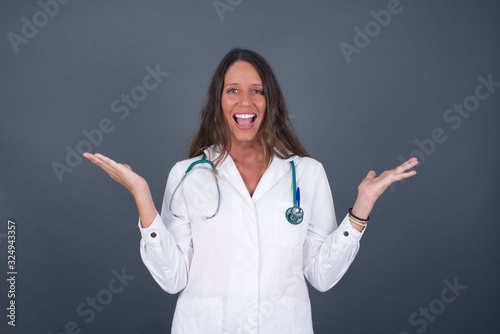 Portrait of beautiful doctor female looks with excitement at camera  keeps hands raised over head  notices something unexpected  isolated over gray wall. Lovely woman reacts on sudden news.