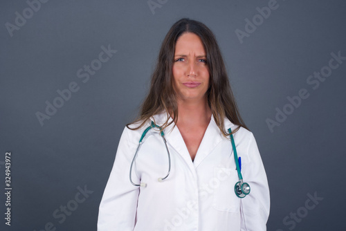 Portrait of outraged young doctor woman wearing medical uniform, frowning her eyebrows being displeased with something. Scowling pretty female isolated over grey.