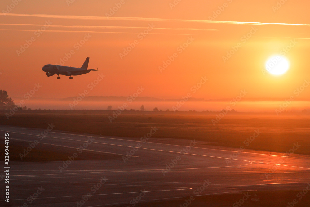 The photo shows a plane landing at dawn or sunset. The crimson rays of the sun nicely illuminate the runway. The photo was taken on a summer fog day at the airport in the Siberian city of Novosibirsk.