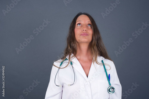 portrait of mysterious charming doctor woman with straight hair looking up with enigmatic smile. Beautiful smiling girl looking up standing against gray wall.