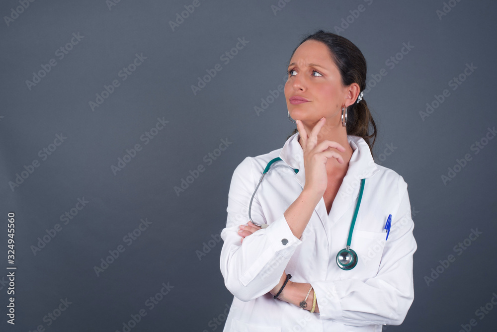 Isolated portrait of stylish young European doctor woman with hand under chin and looking sideways with doubtful and skeptical expression, suspect and doubt. Standing indoors over gray background. Isolated portrait of stylish young European doctor woman with hand under chin and looking sideways with doubtful and skeptical expression, suspect and doubt. Standing indoors over gray background.