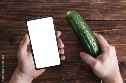 male left hand holding a white screen smartphone on a brown wooden background while in the right cucumber
