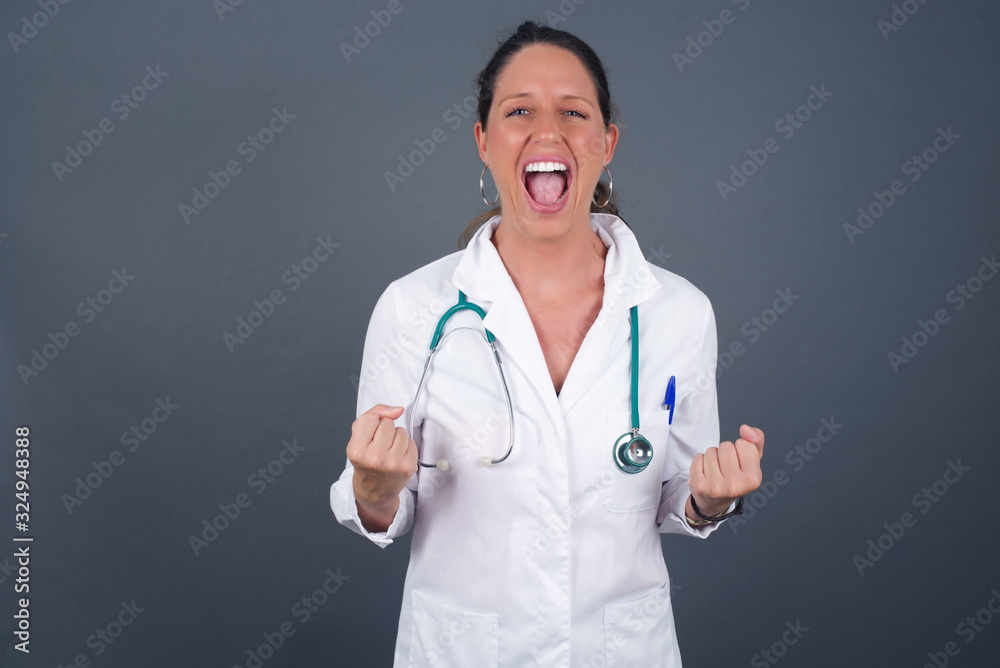 Close up portrait of doctor woman wearing medical uniform raising fists up isolated on shine grey background big eyes and wide open mouth. Close up portrait of doctor woman wearing medical uniform raising fists up isolated on shine grey background big eyes and wide open mouth.