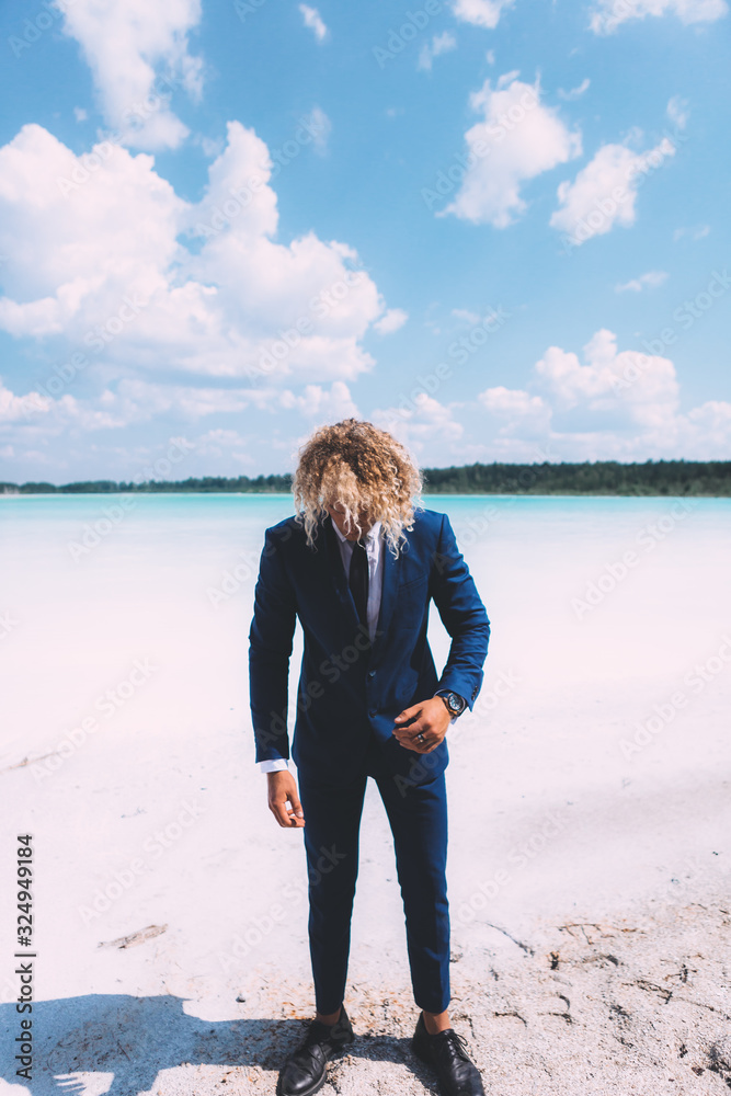 Portrait of blond curly man dressed blue business suit. Clear water and blue sky on the background. Summer time. Travel topic. Portrait of blond curly man dressed blue business suit. Clear water and blue sky on the background. Summer time. Travel topic.