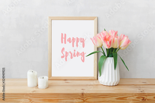 Frame with text HAPPY SPRING, white ceramic vase with bouquet of pink tulips flowers, candles on a wooden table or shelf on a background of light gray wall. Stylish spring home interior decor. Mock up