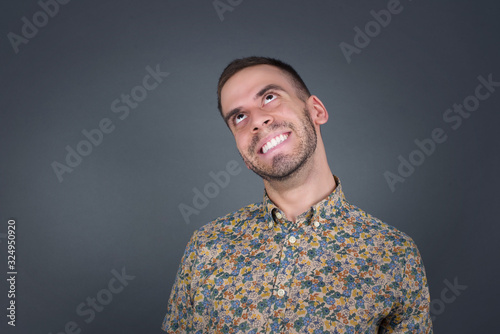 Portrait of mysterious charming blond male looking up with enigmatic smile. Handsome smiling guy looking up standing against gray wall.