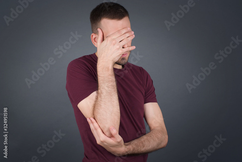 Indoor portrait of handsome man with stylish haircut, wearing casual clothes, making facepalm gesture while smiling, standing over gray background amazed with stupid situation. photo