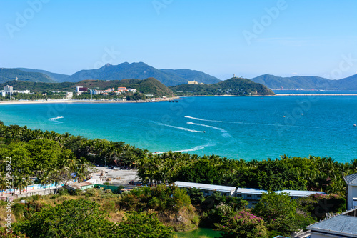 Fototapeta Naklejka Na Ścianę i Meble -  View from the hotel to clear turquoise sea, mountains, beach, jet skis, motor boats and parachutes on the coast of Dadonghai Bay in South China Sea. Sunny day
