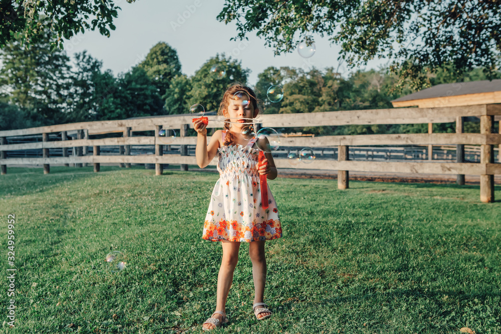 Candid portrait of preschool Caucasian child girl blowing soap bubbles in park at summer sunset. Real authentic happy childhood moment. Lifestyle children outdoor seasonal activities.