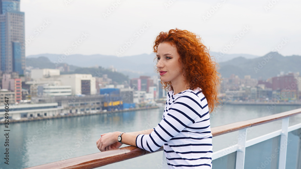 Fototapeta premium Portrait of a curly-haired red-haired girl on the background of high-rise buildings of the city. young beautiful girl on a background of the silhouette of the city posing in life style clothes