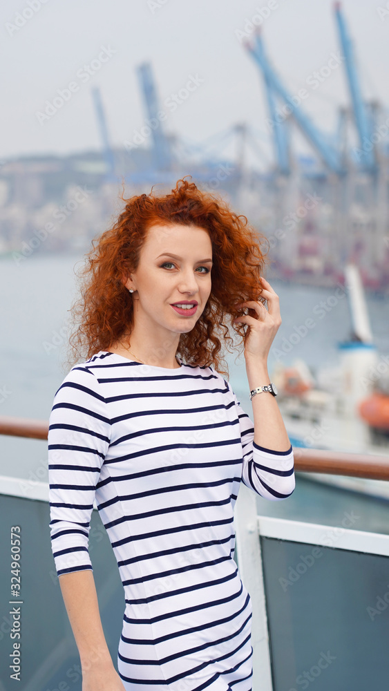 beautiful young red-haired curly girl on the background of port cranes. happy girl posing in a striped dress, emotion, joy, smile. portrait