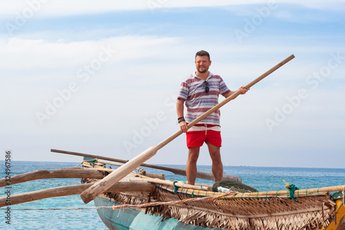 Portrait of a man with a middle beard in a shirt and red shorts on a traditional fisherman boat with a paddle in his hands.