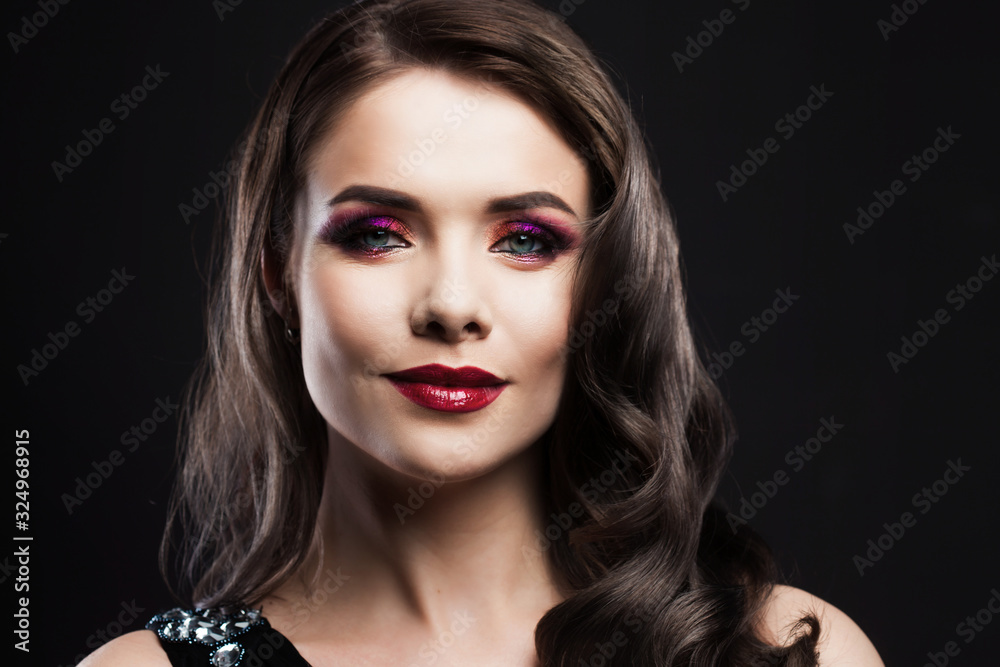 Fototapeta premium Portrait of a young luxurious woman in a low key. Bright makeup with scarlet lipstick, hairstyle with curls,