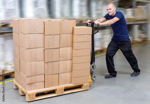 Warehouseman in storehouse fast-moving pallet with stacker. Worker in warehouse moves pallet in rack system. Caucasian labourer working with small forklift. Man rush with pallet jack.