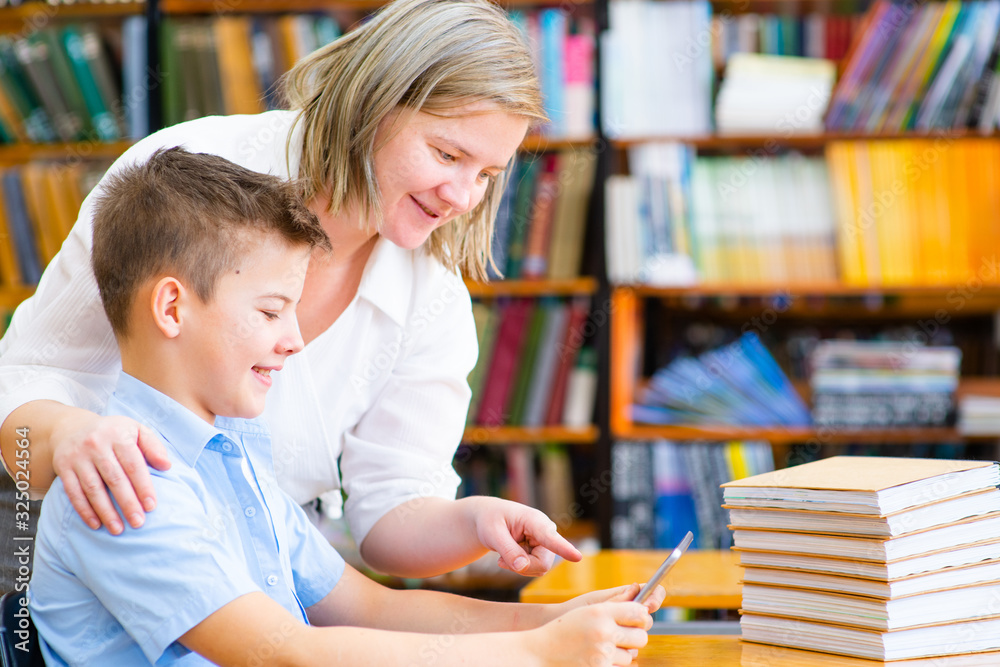 The teacher helps the student in the library. The boy holds a tablet in his hands and looks at it. The teacher shows a finger to the tablet The teacher helps the student in the library. The boy holds a tablet in his hands and looks at it. The teacher shows a finger to the tablet