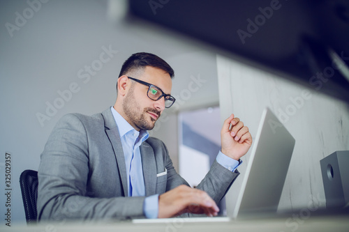 Hardworking classy handsome businessman in suit and with eyeglasses sitting in his office and using laptop.