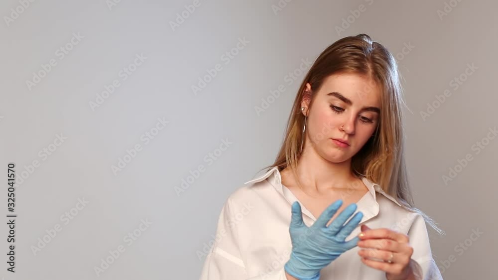 caucasian female with piercing in nose standing, looking at camera in silence, removes blue glove from her hand. young pretty girl in shirt with long blond hair stands against white studio background caucasian female with piercing in nose standing, looking at camera in silence, removes blue glove from her hand. young pretty girl in shirt with long blond hair stands against white studio background