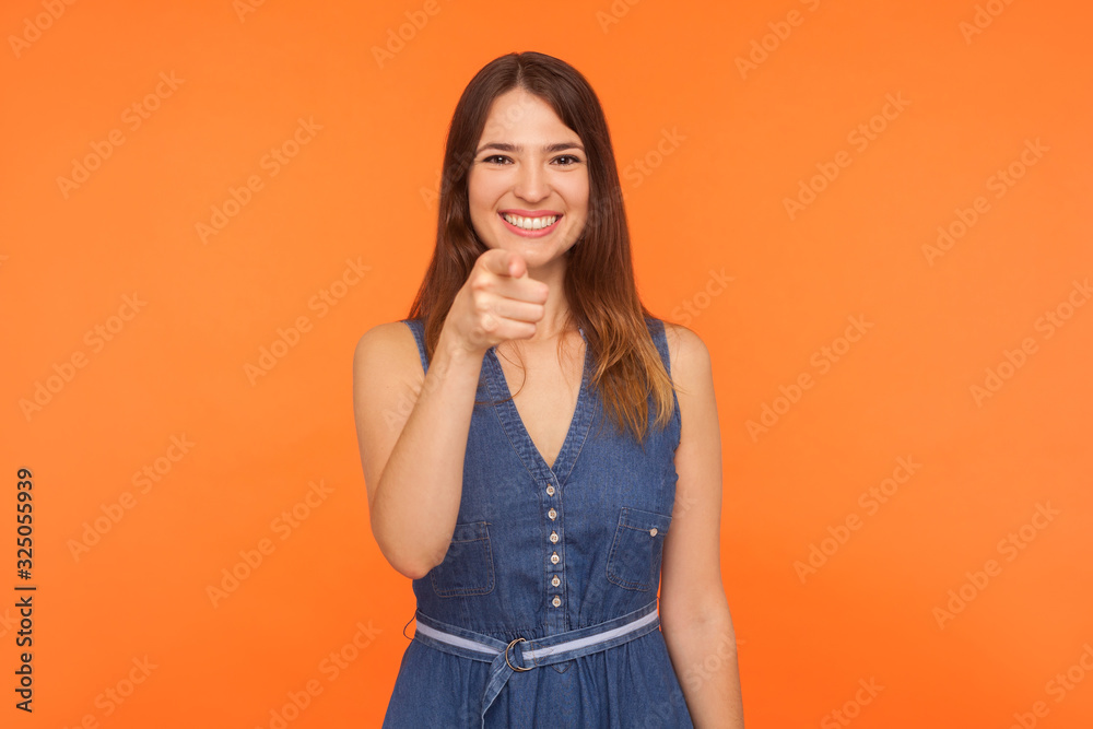Hey you! Positive smiling beautiful brunette woman in denim dress pointing finger to camera, selecting choosing with satisfied glad expression. indoor studio shot isolated on orange background Hey you! Positive smiling beautiful brunette woman in denim dress pointing finger to camera, selecting choosing with satisfied glad expression. indoor studio shot isolated on orange background